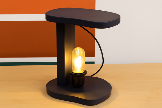 "Modern black C-shaped table lamp with exposed Edison bulb glowing warmly, placed on a light wooden surface with colorful background wall art."