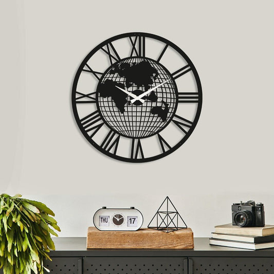 "Black metal wall clock with world map design and Roman numerals, mounted on a neutral-colored wall above a decorative shelf"