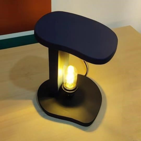 "Modern black C-shaped table lamp with exposed Edison bulb glowing warmly, placed on a light wooden surface with colorful background wall art."