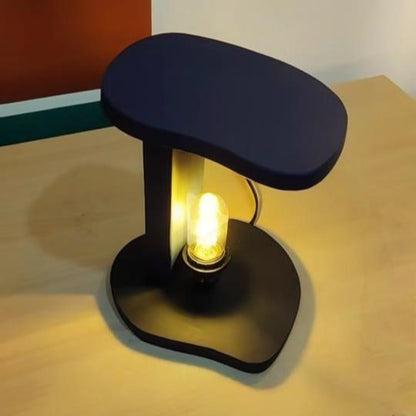 "Modern black C-shaped table lamp with exposed Edison bulb glowing warmly, placed on a light wooden surface with colorful background wall art."