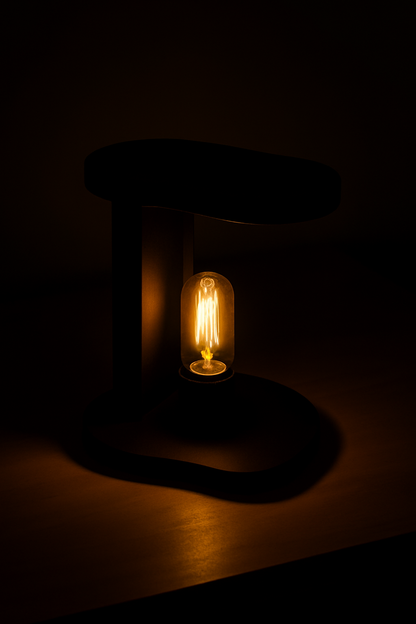 "Modern black C-shaped table lamp with exposed Edison bulb glowing warmly, placed on a light wooden surface with colorful background wall art."