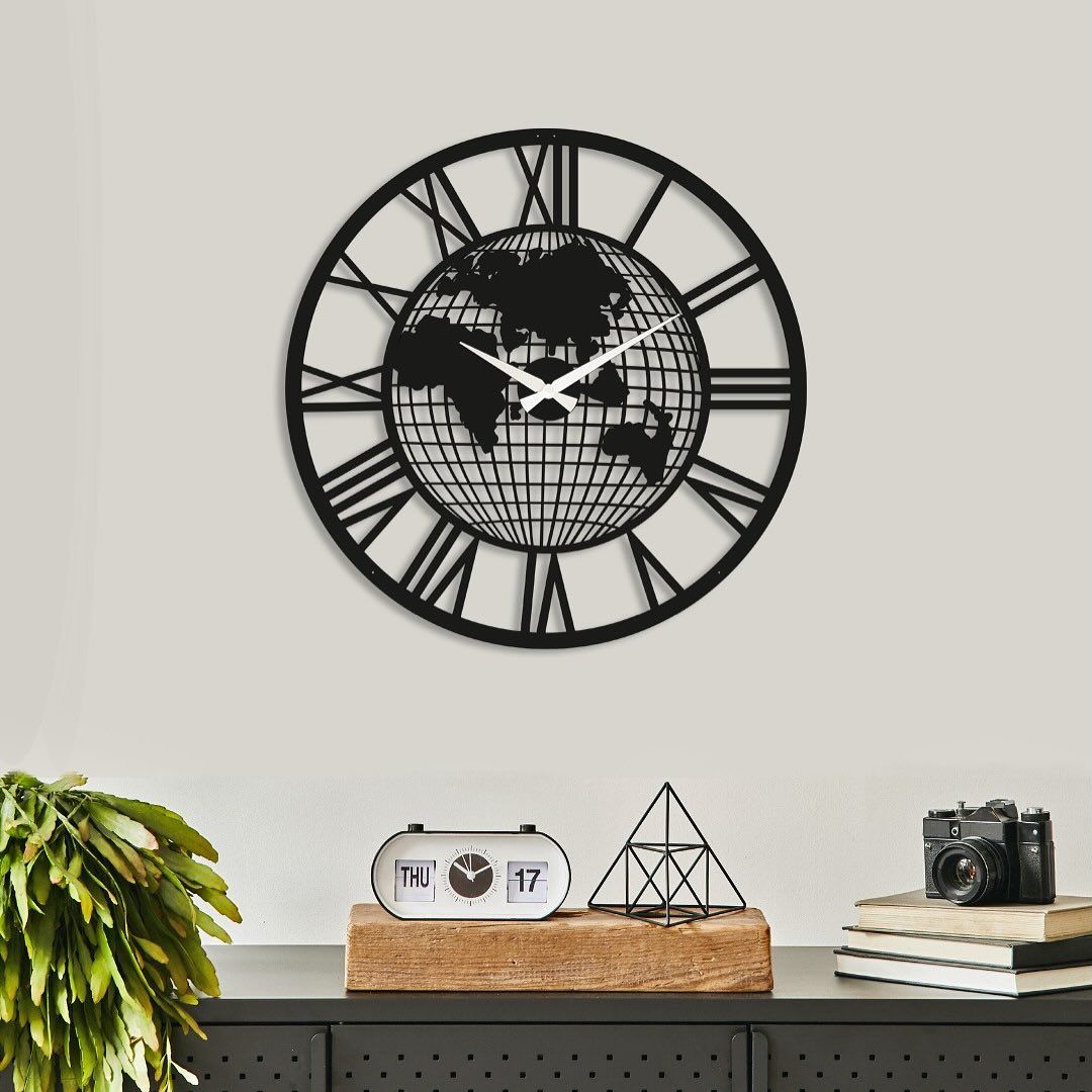 "Black metal wall clock with world map design and Roman numerals, mounted on a neutral-colored wall above a decorative shelf"